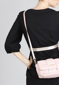 Růžová kožená crossbody kabelka s prošívaným designem, zlatými doplňky a dlouhým popruhem, nošená přes černé šaty s nabíranými rukávy.