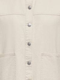 Vêtement couleur crème avec boutons-pression métalliques et deux poches plaquées à l'avant, montrant des coutures détaillées et la texture du tissu.