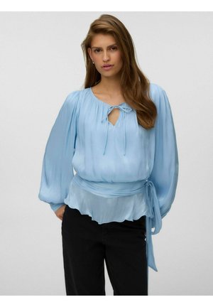 Femme aux longs cheveux bruns porte un chemisier bleu clair à manches longues avec un col à nouer et une ceinture à la taille, associé à un pantalon noir.