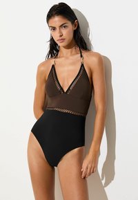 Nedves sötét hajú nő fekete és barna egyrészes fürdőruhában, hálós részletekkel, fehér fal előtt áll napfényben.