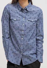 Blå långärmad skjorta med knappar och mörka små abstrakta mönster, två bröstfickor med lock, bärs otam.