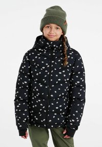 Zwarte pufferjas met een wit bloemenpatroon, vol rits, hoge kraag en capuchon; gecombineerd met een groene gebreide muts. Ook groene broek is zichtbaar.
