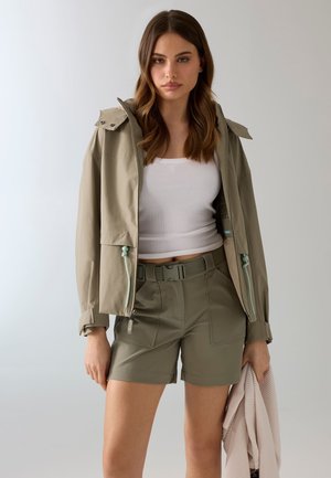 Mujer con cabello castaño ondulado, lleva una chaqueta beige, top blanco sin mangas, shorts verdes oliva con cinturón, sosteniendo un suéter rosa claro doblado.