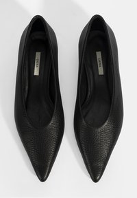 Escarpins noirs en cuir à bout pointu avec une finition texturée, présentant un intérieur lisse et un design minimaliste. Étiquette de la marque visible à l'intérieur.