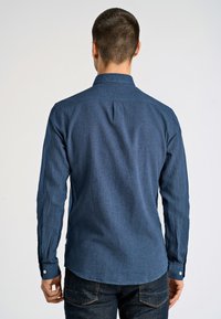 Man in een donkerblauwe lange mouwen shirt en donkere jeans, met de rug naar de camera voor een lichte effen achtergrond.