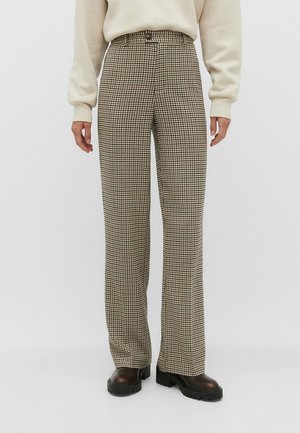 Person iført beige og brune højttaljede bukser med højtaljemønster i hundetand, sorte chunky støvler og en cremefarvet sweater.