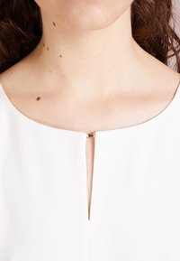 Crop top blanc avec un col rond et un petit accent en métal doré au centre. Tissu lisse avec une texture minimale.