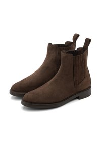 Une paire de bottes Chelsea en daim marron foncé avec des panneaux élastiques sur les côtés et des languettes de tirage sur un fond blanc.