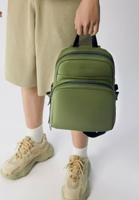 Grön faux läder ryggsäck med två dragkedja fack, svarta detaljer och ett handtag på toppen, bärs av en person i shorts och sneakers.