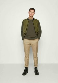 Oliven grønn uten ermer jakke over en mørkegrønn ribbestrikket genser, kombinert med lysebeige avsmalnende bukser og svarte ankelboots.