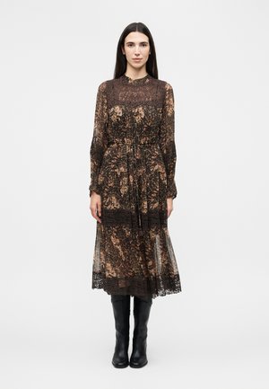 Žena stojaca čelom vpred, oblečená v dlhý rukáv hnedé kvetinové šaty s čipkovými detailmi a čiernymi čižmami po kolená na bielom pozadí.