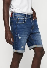 Denim kratke hlače v temno modri barvi z unikatnimi podrobnostmi, zavihki na robovih in zlato nitjo. Imajo sprednje žepke in zapiranje na gumb.