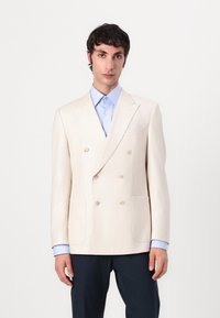 Double-breasted cream blazer made of textured fabric, featuring two front pockets, notched lapels, and cream buttons; worn over a light blue shirt.