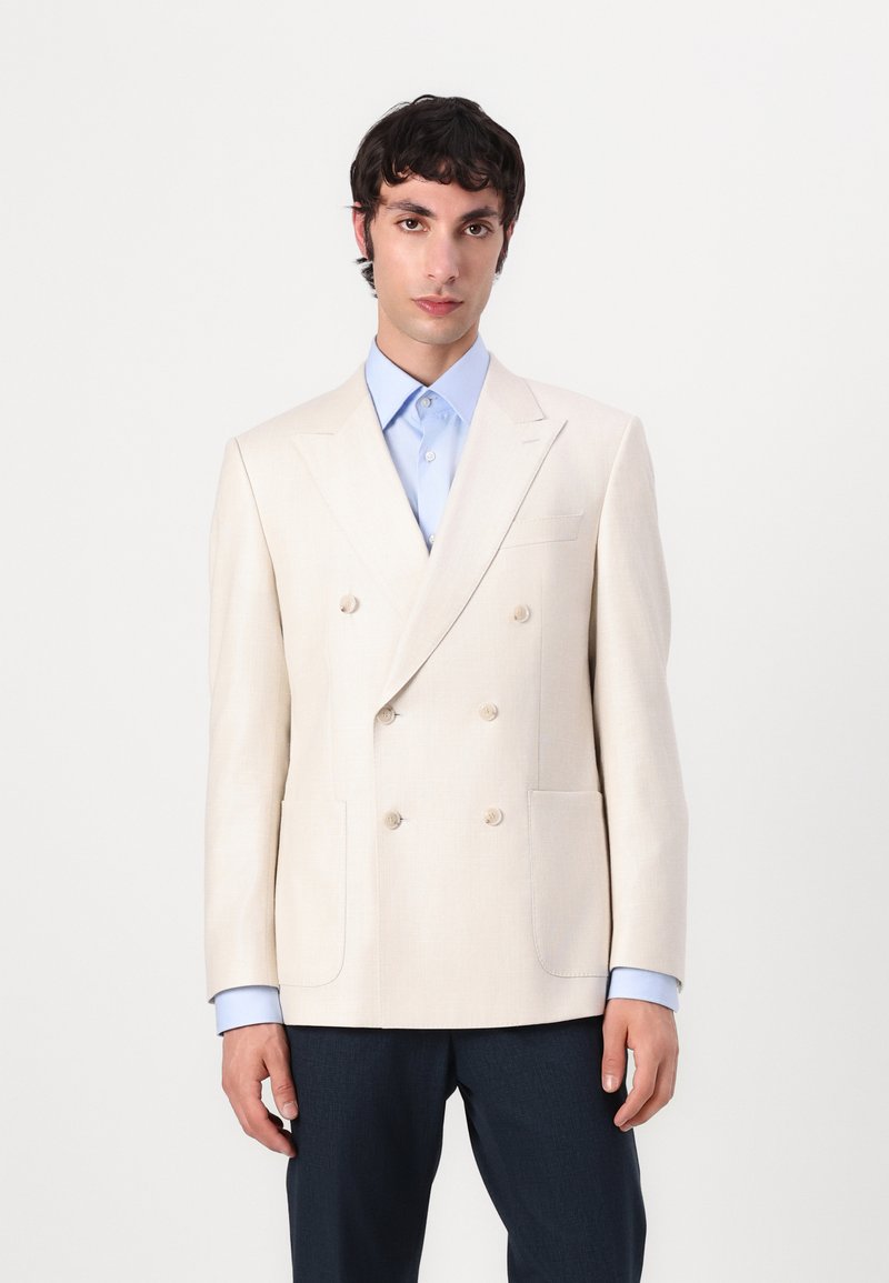 Double-breasted cream blazer made of textured fabric, featuring two front pockets, notched lapels, and cream buttons; worn over a light blue shirt.