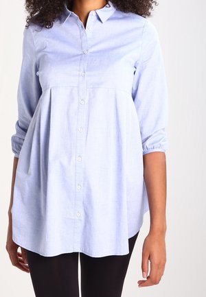 Femme portant un chemisier bleu clair à manches longues avec boutons et une taille ample froncée, associé à un pantalon noir.
