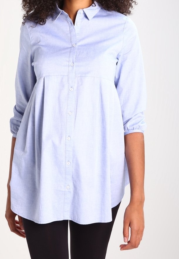 Femme portant un chemisier bleu clair à manches longues avec boutons et une taille ample froncée, associé à un pantalon noir.