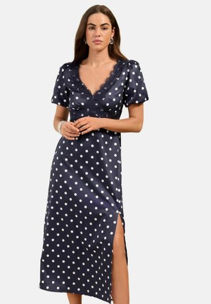 Žena v tmavomodrom midi šatách s krátkym rukávom, bodkami, čipkovaným výstrihom do V a bočným rozparkom, stojaca pred jednoduchým pozadím.