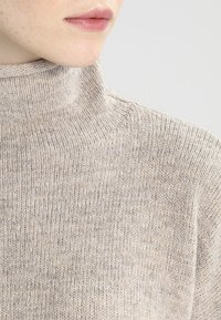 Nahaufnahme des Untergesichts und des Halses einer Person, die einen beigen gestrickten Rollkragenpullover vor einem einfarbigen Hintergrund trägt.