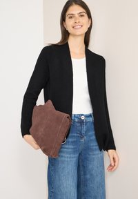 Czarny prążkowany sweter na guziki noszony na białej koszuli, w połączeniu z szerokimi niebieskimi dżinsami, trzymający brązową zamszową kopertówkę.