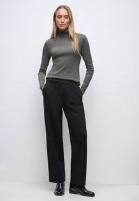 Maglione a collo alto verde oliva abbinato a pantaloni neri a gamba larga. Il modello sta in piedi con le mani in tasca, indossando stivali neri alla caviglia. Design semplice.