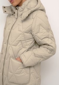 Bēšas krāsas quilted jaka ar kapuci. Iekļauj ģeometrisko šuvumu, rāvējslēdzēja aizdari un lielas priekšējās kabatas. Mīksta tekstūra, minimāla apdare.