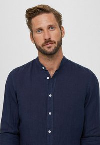 Camicia in lino blu navy a maniche lunghe con apertura frontale a bottoni, caratterizzata da un colletto a banda e una trama sottile. Cinque bottoni bianchi visibili.