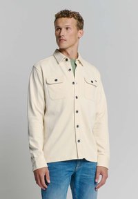 Lichtcrème hemdjacket met twee borstzakken, zwarte knopen en een rechte pasvorm. Gedirigeerd boven een groene top en gecombineerd met blauwe spijkerbroek.