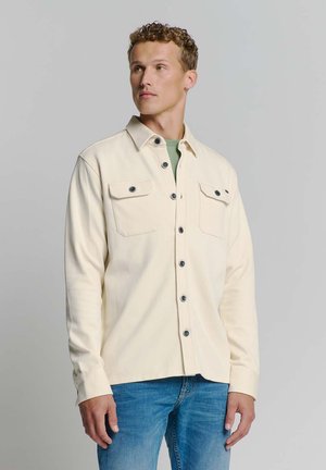 Light cream shirt jacket with two chest pockets, black buttons, and a straight fit. Worn over a green top and paired with blue jeans.