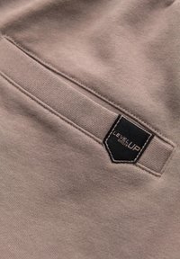 Gros plan d’un tissu beige avec une poche passepoilée cousue et une étiquette noire portant l’inscription « Level Up » sur le bord supérieur.