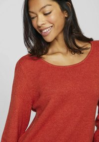 Giovane donna con tono di pelle medio e capelli scuri sorride dolcemente, indossando un maglione a maglia color ruggine con un ampio scollo.