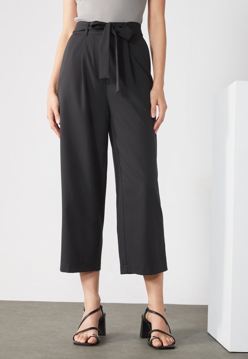 Pantalons larges noirs avec une ceinture nouée à la taille, présentant une texture de tissu lisse et une longueur courte, associés à des talons noirs à lanières.