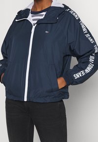 Person wearing navy blue zip-up jacket with white zipper, "Tommy Jeans" text on sleeves, and hands in pockets, paired with black jeans.