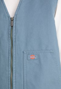 Gilet en toile bleue avec une fermeture à glissière centrale en métal, doté d'un logo en patch sur la poche avant gauche et d'une surface en tissu texturé.