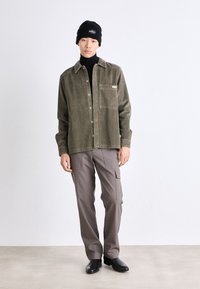 Olijfgroene corduroy shirtjas met een voorkeep, gecombineerd met lichtbruine cargo-broeken en zwarte enkellaarzen. Zwarte gebreide beanie inbegrepen.