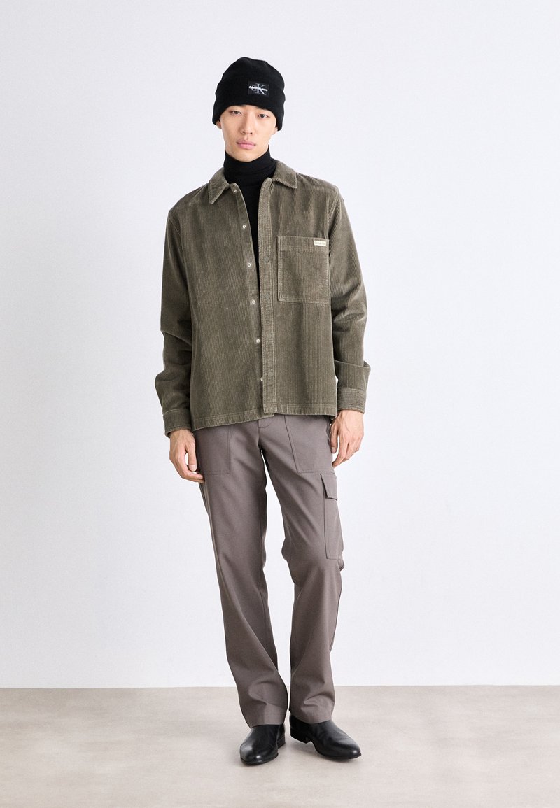 Oliwkowa koszula kurtka z sztruksu z kieszenią z przodu, zestawiona z jasnobrązowymi spodniami cargo i czarnymi skórzanymi botkami. W zestawie czarna czapka dzianinowa.