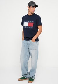 Mørkeblå bomuld t-shirt med trykt logo, lyseblå baggy jeans og grønne sneakers med brune såler. Kappen matcher skjortefarven.