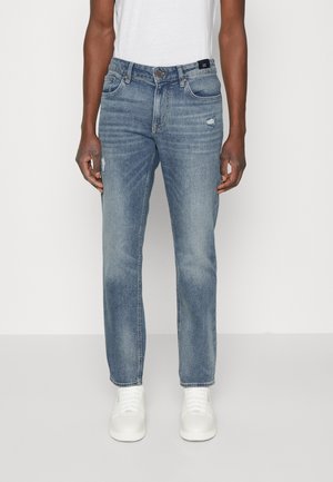 Uomo che indossa jeans azzurri slavati a gamba dritta con lievi segni di usura sulle cosce e sneakers bianche, in piedi davanti a uno sfondo bianco.