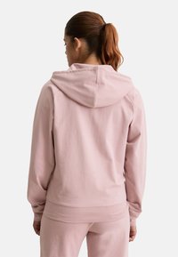 Femme aux cheveux roux attachés en queue de cheval, portant un sweat à capuche rose clair et un pantalon assorti, tournée le dos devant un fond blanc uni.