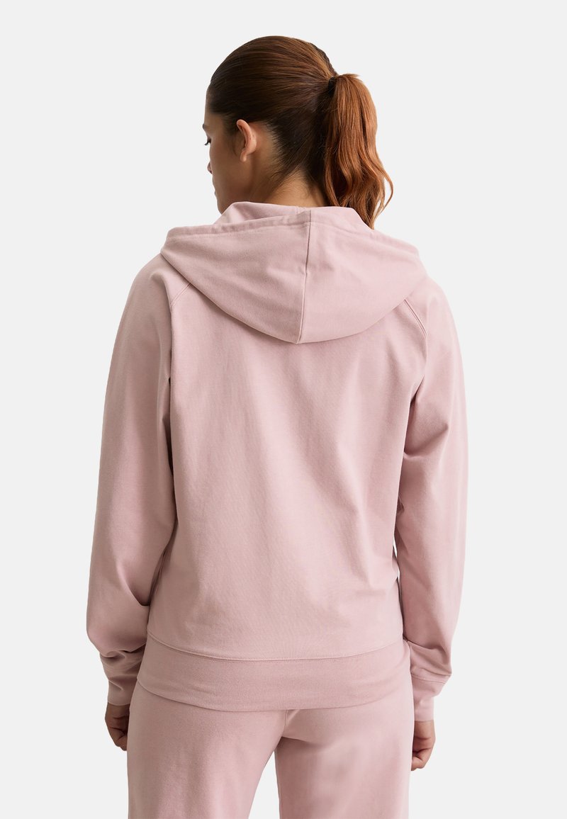 Vrouw met kastanjebruin haar in een paardenstaart, gekleed in een lichtroze hoodie en bijpassende broek, met haar rug naar de camera gekeerd tegen een eenvoudige witte achtergrond.
