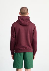 Vínová mikina s kapucí s velkou zadní kapsou a volným střihem, doplněná zelenými šortkami. Má silné manžety, strukturovanou látku a prostornou kapuci.
