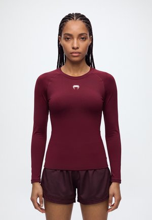 Une femme aux cheveux tressés se tient face à l’objectif, vêtue d’un haut de sport bordeaux à manches longues et d’un short assorti, devant un fond uni clair.