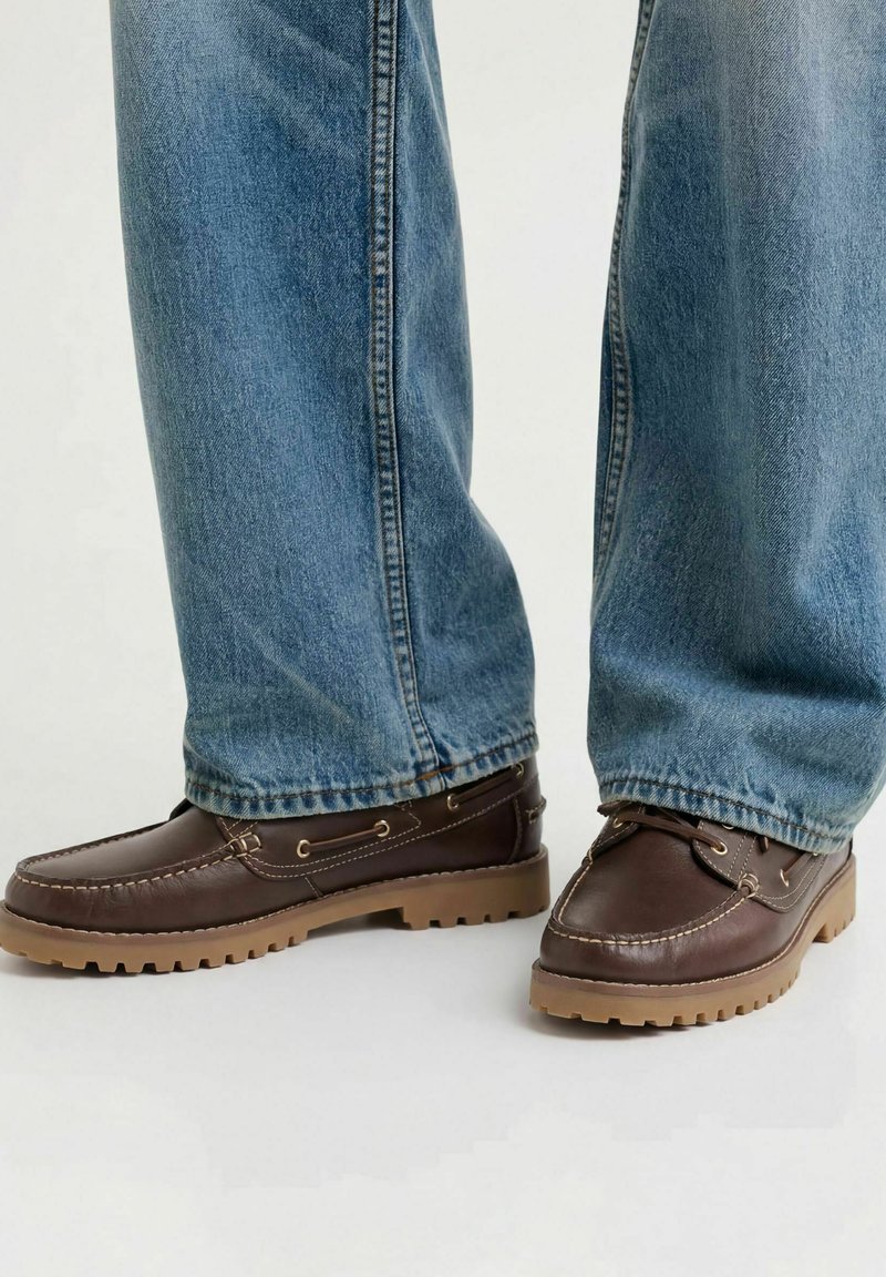 Personne portant des mocassins en cuir marron avec des semelles couleur beige et un jean en denim bleu, debout sur une surface claire.