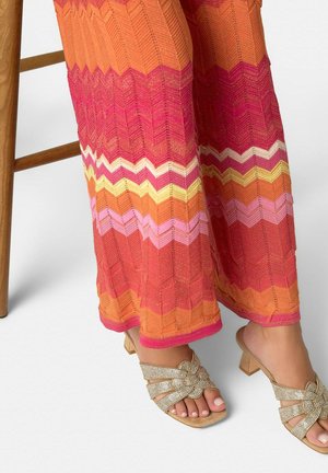 Piernas con pantalones anchos de patrón de zigzag colorido y sandalias de tacón beige con brillo, descansando cerca de la pata de una silla de madera.