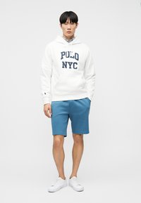 Άνδρας που φοράει λευκή μπλούζα με κουκούλα με το κείμενο "POLO NYC", μπλε σορτς και λευκά αθλητικά παπούτσια, στέκεται μπροστά σε μια απλή λευκή επιφάνεια.