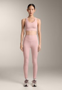 Femeie stând în picioare, purtând un top sportiv și colanți asortați de culoare roz deschis, cu adidași bej și albi, pe un fundal neutru.