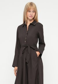 Braunes Hemdblusenkleid mit langen Ärmeln, Kragen, Taillengurt und seitlichen Taschen. Der glatte Stoff und der lockere Schnitt betonen das Design.