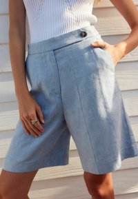 Shorts en lin bleu clair avec une coupe ajustée, dotés d'une fermeture à un bouton, de poches latérales et de plis à l'avant. Texture lisse.