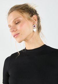 Silberblomster øreringer med hjerteform, som har en lilla edelsten og teksturerte kronblade, båret sammen med en svart ribbet topp.