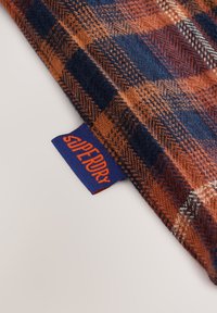 Rutet stof i orange og marineblå med en blød tekstur. Har en synlig blå label med "Superdry" i orange bogstaver.