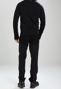 Man står med ryggen mot kameran, klädd i svart utomhusjacka, svarta taktiska byxor och svarta vandringskängor mot en ljus enfärgad bakgrund.